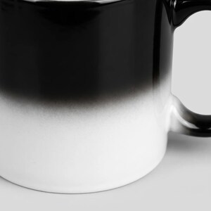 May include: A ceramic mug featuring a smooth gradient transition from a solid black top to a crisp white base. The cylindrical vessel includes a curved handle on the right side, showcasing a clean, minimalist aesthetic with a seamless color blend.