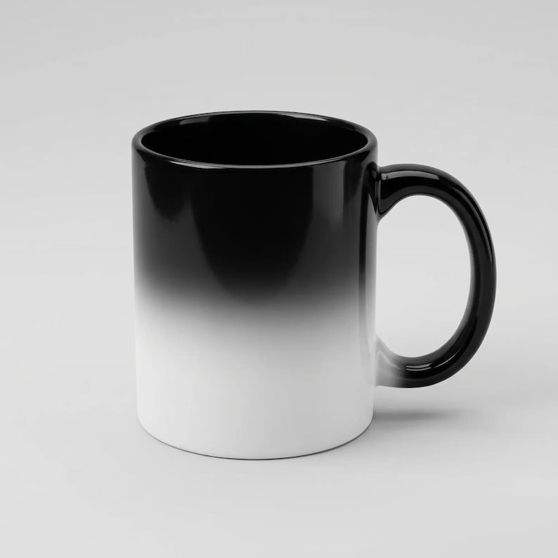 Puede incluir: Una taza de cer&aacute;mica con un degradado de color suave que transiciona desde un negro s&oacute;lido en el borde superior y el asa hasta un blanco n&iacute;tido en la base. El recipiente cil&iacute;ndrico tiene un asa curva en el lado derecho y un acabado brillante.
