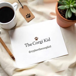 Puede incluir: Una tarjeta rectangular blanca con el texto negro The Corgi Kid y el usuario de redes sociales @cypherthecorgikid, acompañado de un pequeño icono de huella marrón. La tarjeta está situada cerca de una taza de café blanca, una etiqueta marrón y una suculenta.