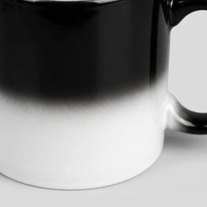 Puede incluir: Una taza de cer&aacute;mica que presenta una transici&oacute;n de degradado suave desde una parte superior negra s&oacute;lida hasta una base blanca n&iacute;tida. El recipiente cil&iacute;ndrico incluye un asa curva en el lado derecho, mostrando una est&eacute;tica limpia con una mezcla de colores.