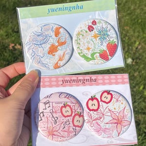 May include: Two clear plastic packages each containing two circular pinback buttons. The top package features goldfish and strawberry designs with the text yueningnha. The bottom package displays apple and floral patterns with the text yueningnha.