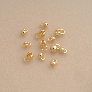 May include: A collection of small, shiny gold-toned metal crimp covers, each featuring a spherical bead shape with an attached loop. These jewelry findings are scattered across the frame, displaying a polished, reflective surface finish.