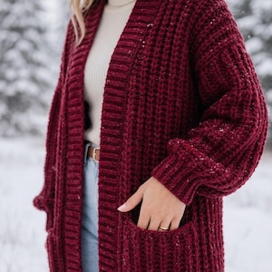 May include: A deep burgundy open-front cardigan featuring a chunky knit texture, long sleeves with ribbed cuffs, and two front patch pockets. The garment is worn over a cream-coloured ribbed turtleneck top and light blue denim jeans with a brown belt.