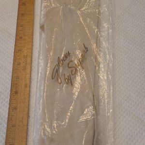 Peut inclure: Une paire de gants blancs scellée dans un sac en plastique transparent avec le texte Gloves by Superb imprimé en doré. Une règle en bois Westcott est placée à côté de l'emballage, montrant une longueur d'environ 28 centimètres.