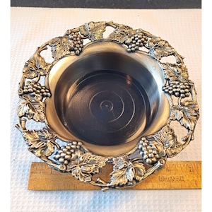 May include: A round silver-toned metal bowl featuring an ornate openwork rim decorated with embossed grape clusters and vine leaves. A wooden ruler placed underneath shows the bowl has a diameter of approximately 6 inches.