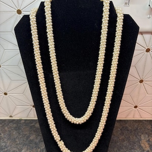 May include: A long, multi-strand necklace composed of numerous small, cream-colored spherical beads arranged in a twisted, rope-like pattern. The necklace features a continuous, uniform texture created by the densely packed, round, off-white beads.
