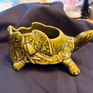 May include: A glossy olive green ceramic planter shaped like a turtle. The shell features a textured relief pattern of overlapping leaves and geometric shapes. The turtle has a raised head and four short legs, with an open cavity on the back.