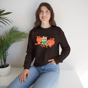 Op de afbeelding: Een donkerbruin sweatshirt met lange mouwen en een grafische print van een wasbeer die een groen boek leest, omringd door oranje pompoenen, herfstbladeren en een mok met slagroom. Het sweatshirt heeft een ronde hals en boorden.