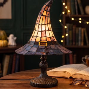 May include: A stained glass table lamp shaped like a pointed witch hat. The top section features amber and orange glass panels, while the brim is made of dark purple and blue glass. It sits on a decorative, dark bronze-colored metal base.
