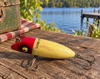 May include: A vintage fishing lure with a red head and a yellow body featuring the text 'BANG-O-LURE' in black lettering. The lure has a gold-colored eye, a metal line tie at the front, and two silver-toned treble hooks attached to the underside.