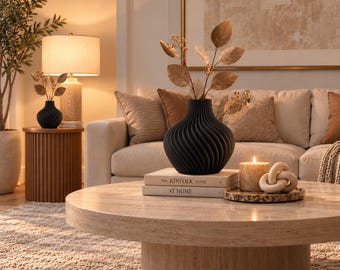 May include: A matte black vase with a spiraled, ribbed texture holds a branch with brown leaves. The vase sits on two stacked books titled The Kinfolk Home and At Home, next to a lit candle and a decorative stone knot on a circular tray.