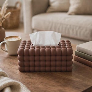 May include: A rectangular tissue box cover featuring a matte brown finish and a repeating bubble-like spherical texture across all visible sides. The cover is designed with a central rectangular opening at the top, through which white tissues emerge.