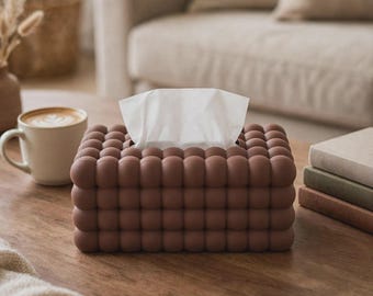 Puede incluir: Una funda rectangular para caja de pañuelos con un acabado marrón mate y una textura esférica repetitiva similar a burbujas en todos los lados visibles. La funda tiene una abertura rectangular central en la parte superior para los pañuelos.