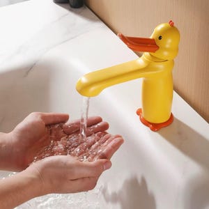 May include: A yellow duck-shaped bathroom faucet with an orange beak and orange feet at the base. The faucet is mounted on a white sink and is actively dispensing a steady stream of clear water into a pair of cupped hands positioned below.