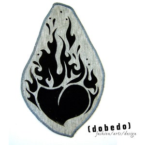 Könnte beinhalten: Ein schwarzer und grauer Stoff-Patch mit einer schwarzen Herzform, die von Flammen umgeben ist. Der Patch hat einen grauen Hintergrund und einen gezackten Rand. Der Text (dobe do) fashion/arts/design ist unter dem Patch gedruckt.
