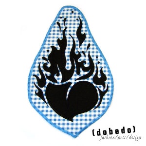 Könnte beinhalten: Schwarz-weiß gestickter Patch mit einer Herzform, die von Flammen umgeben ist, auf einem blau-weiß karierten Hintergrund. Der Text "(dobe do)" befindet sich unter dem Patch. "fashion/arts/design" befindet sich unter dem Text.