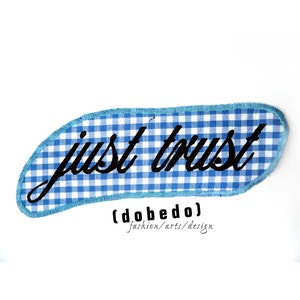 Könnte beinhalten: Ein blauer und weißer Gingham-Stoff-Patch mit dem Text "just trust" in schwarzer kursivschrift. Der Patch hat einen blauen gestickten Rand und der Text "(dobedo) fashion/arts/design" ist unter dem Patch gedruckt.