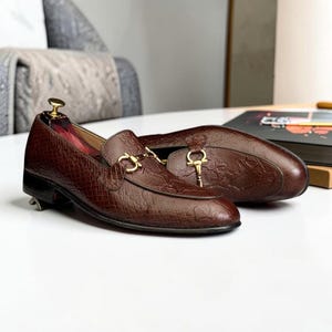 May include: A pair of brown loafers featuring a textured snakeskin-like pattern and a gold-toned metal horsebit detail across the vamp. The shoes have a low heel and a rounded toe, with a wooden shoe tree inserted into the front of the shoe.