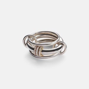 Könnte beinhalten: Ein mehrreihiger Ring aus mehreren ineinandergreifenden silberfarbenen Metallschleifen. Drei der mittleren Ringe sind mit kleinen, klaren, funkelnden Steinen besetzt, die in goldfarbenes Metall gefasst sind und einen Kontrast zur glatten Silberoberfläche bilden.