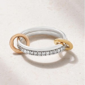 May include: A silver ring featuring a band encrusted with a row of small clear stones. Two smooth, circular gold-toned links are attached to the main band, one on each side, creating a layered appearance with contrasting metallic finishes.