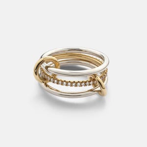 Könnte beinhalten: Ein mehrreihiger Ring aus polierten silber- und goldfarbenen Metallbändern. Das Design umfasst ein mittleres Band, das mit kleinen, klaren, funkelnden Steinen besetzt ist, sowie zwei kleinere goldfarbene Ringe, die mit den Hauptbändern verbunden sind.