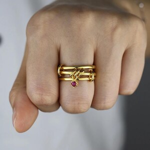 Puede incluir: Un anillo dorado de varias bandas que presenta tres aros circulares lisos y superpuestos, conectados por un pequeño dije colgante. El dije consiste en una sola piedra preciosa redonda de color rojo rosado vibrante.