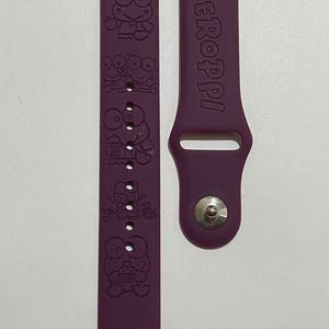 May include: A purple silicone watch strap featuring embossed Keroppi character graphics on one side and the text Keroppi embossed on the other. The strap includes a silver-toned metal pin fastening and multiple adjustment holes for a secure fit.