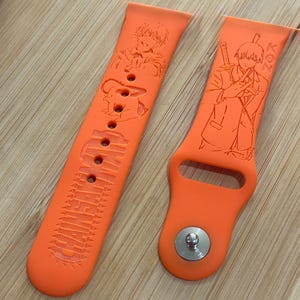 May include: A bright orange silicone watch strap featuring engraved anime-style character illustrations and the text "CHAINSAW MAN". The strap includes a metallic silver pin closure and multiple adjustment holes for a secure fit on the wrist.