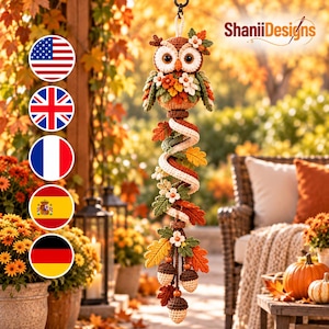 May include: A hanging crochet decoration featuring a brown owl with large eyes, adorned with autumn-coloured leaves, small white flowers, and three dangling acorns. The piece includes a spiral element and the text ShaniiDesigns.