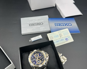 May include: A Seiko Velatura chronograph watch with a blue dial, gold-tone bezel, and silver-tone metal link band, presented in a black box with a white Seiko branded box, instruction manual, warranty card, and a small Seiko Velatura tag.