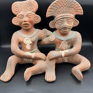 Puede incluir: Una escultura de arcilla color terracota que representa dos figuras sentadas y unidas. La figura de la izquierda tiene el cabello en forma de espiral, mientras que la de la derecha lleva un gran tocado en forma de abanico. Ambas tienen collares.