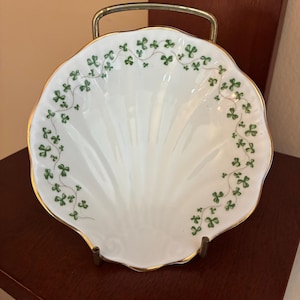May include: A white porcelain shell-shaped dish featuring a delicate green shamrock vine pattern along the scalloped edge. The dish has a thin gold trim around the rim and is displayed on a simple gold-toned metal stand for upright viewing.