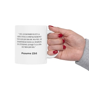 May include: A white ceramic mug featuring black printed text in French that reads, "OUI, LE BONHEUR ET LA GRÂCE M'ACCOMPAGNERONT TOUS LES JOURS DE MA VIE, ET J'HABITERAI DANS LA MAISON DE L'ÉTERNEL JUSQU'À LA FIN DE MES JOURS." followed by "Psaume 23:6".