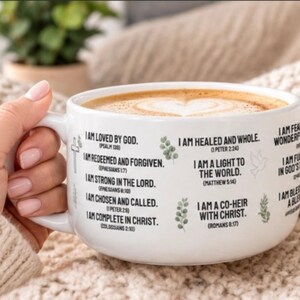 以下が含まれることがあります： ハート型のフォームデザインが施されたコーヒー入りの白いセラミックマグカップ。外側には「I AM LOVED BY GOD」「I AM HEALED AND WHOLE」「I AM A LIGHT TO THE WORLD」などの黒いテキストと、小さな緑色の葉のアイコンがプリントされています。