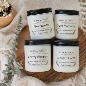 May include: Four white jars with black lids, each featuring a label for Rebekah's Nest hand-crafted goods. The labels are titled Evergreen, Quiet Moments, Cherry Blossom, and Autumn Home, listing organic tallow and beeswax as ingredients.