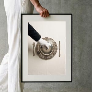 Puede incluir: Una impresión de arte mural enmarcada que muestra una vista cenital de una campana de plata siendo levantada por una mano con guante blanco. Un tenedor y un cuchillo de plata están colocados sobre un mantel blanco a ambos lados del plato.