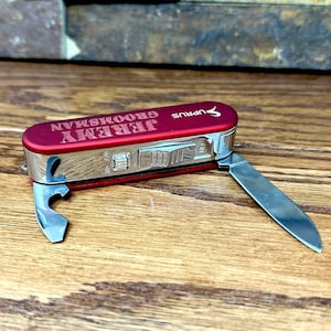 May include: A red pocket knife with a bottle opener and a small blade. The knife is engraved with the text "Jeremy  Groomisman  Army".