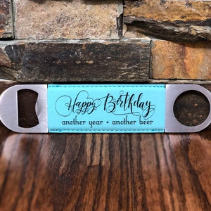 Puede incluir: Un abridor de botellas de cuero azul con un abridor de metal plateado en cada extremo. La parte de cuero tiene el texto "Happy Birthday another year another beer" en negro.
