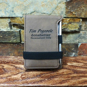 May include: A brown leather notebook with a black elastic band and a silver pen. The notebook is personalized with the text "Tim Pogorelc donebetter Personalized Gifts".