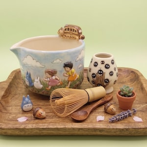 May include: A ceramic matcha bowl featuring a painted scene of two children and two small creatures running through a field, with a cat-shaped bus figurine on the rim. Includes a bamboo whisk, a wooden spoon, a small cup with soot sprites, and two acorn figurines.