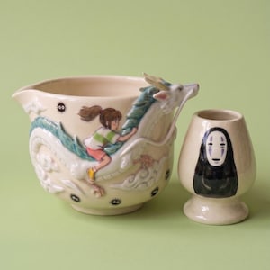 Può includere: Una brocca in ceramica color crema con un rilievo di un personaggio che cavalca un lungo drago dalle scaglie blu, accentuato da piccoli spiritelli della fuliggine neri. Accanto si trova una tazza in ceramica color crema abbinata con una figura nera.