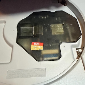 Peut inclure: Un appareil électronique gris clair avec un compartiment ouvert contenant un couvercle noir translucide et une carte microSD SanDisk Extreme 256GB rouge et or. Une étiquette d'avertissement indique NE JAMAIS TOUCHER LA LENTILLE.