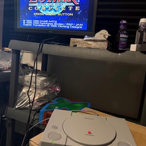 Può includere: Una console di gioco Sony PlayStation grigia con coperchio del disco circolare, pulsante di accensione e porte per controller. Lo schermo mostra il titolo Lunar Silver Star Story Complete con grafica spaziale blu e testo bianco sul copyright.
