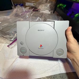 May include: A gray Sony PlayStation game console featuring a circular disc cover, a power button, a reset button, and an open button. The console displays the Sony logo at the top and the colorful PlayStation logo in the center of the front panel.