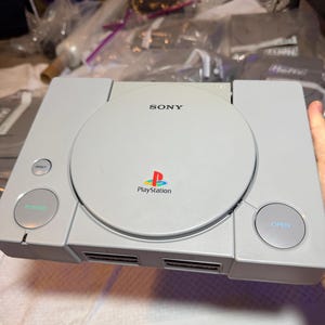 May include: A light gray Sony PlayStation game console featuring a circular disc cover with the Sony and PlayStation logos in the center. The console includes a power button, a reset button, and an open button, along with two controller ports.