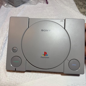 Può includere: Una console di gioco Sony PlayStation grigia dotata di un coperchio circolare per il disco al centro con il logo Sony sopra l'emblema PlayStation. La console include un pulsante di accensione circolare, un tasto reset e un tasto open sul pannello.