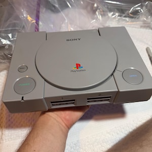 Puede incluir: Una consola de videojuegos Sony PlayStation gris que presenta una cubierta de disco circular con el logotipo de PlayStation, un botón de encendido, un botón de reinicio, un botón de apertura y dos puertos de mando etiquetados 1 y 2 al frente.
