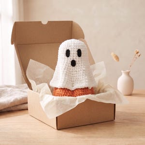 Puede incluir: Un juguete de peluche de fantasma tejido a ganchillo en color blanco con ojos ovalados negros y una boca redonda, colocado dentro de una caja de regalo de cartón marrón abierta con papel de seda blanco. El fantasma tiene una base naranja.