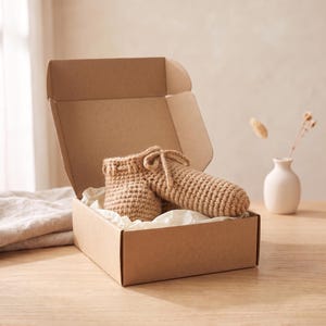 Op de afbeelding: Een beige gehaakt fallisch object met een trekkoord aan de bovenkant, geplaatst in een open bruine kartonnen verzenddoos bekleed met wit vloeipapier. Het item heeft een getextureerde, met de hand gestikte garenconstructie.