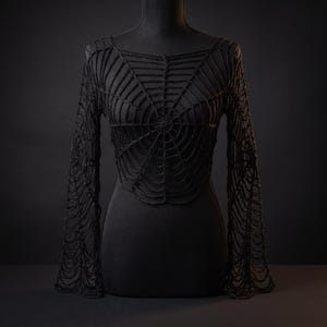 May include: A black long-sleeved top featuring an intricate, open-knit crochet pattern that mimics the design of a spider web. The garment has a wide neckline and fitted sleeves that extend to the wrists, showcasing a detailed, textured yarn construction.